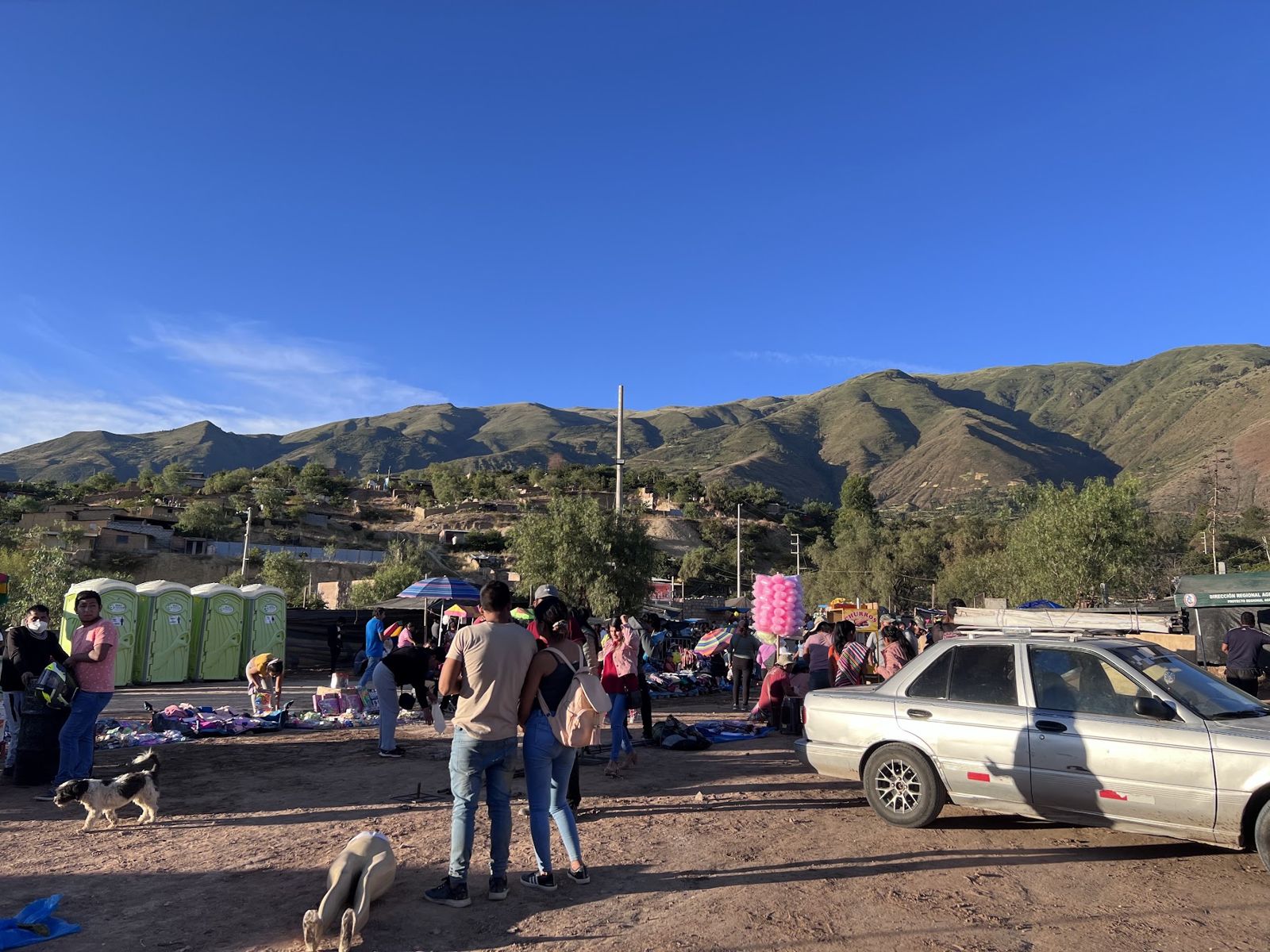 A fairground on open ground below green Andean hills, with stalls and people walking