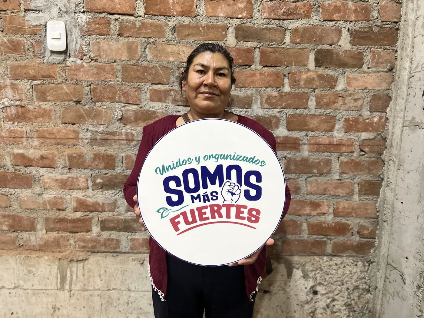 A woman stands against a brick wall holding a round campaign sign
