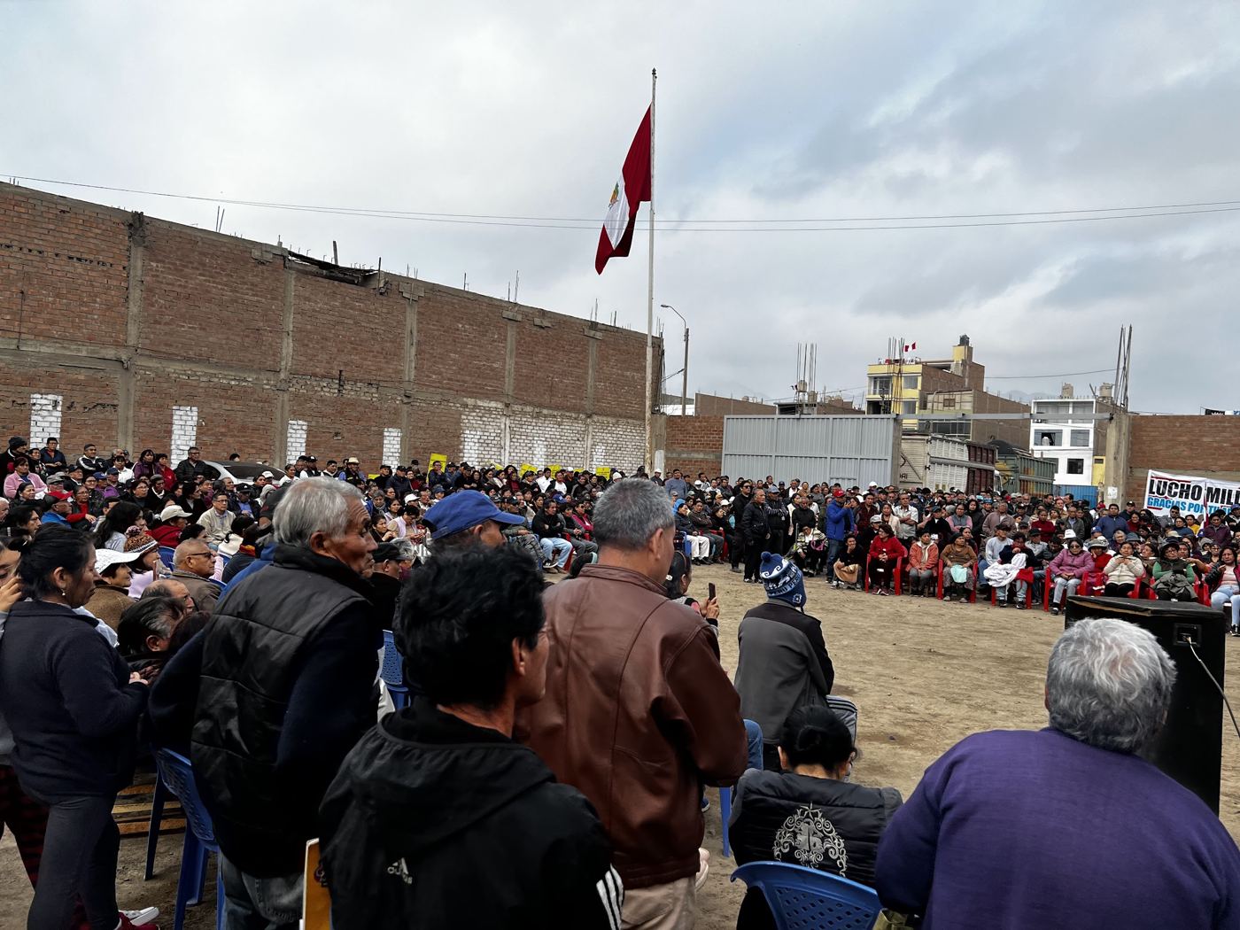 A large community assembly gathered on open ground under a Peruvian flag